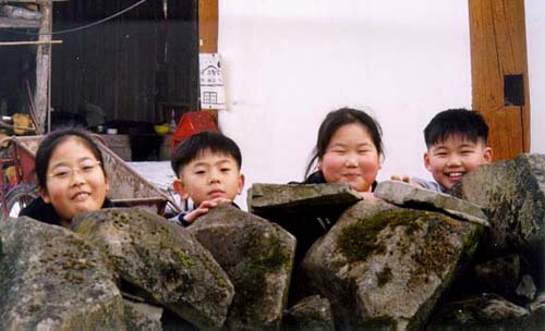 돌담에 얼굴을 내밀고 있는 아이들. 아이들이 못 볼 것을 보았는지 돌담에 얼굴을 내밀며 환하게 웃고 있다 